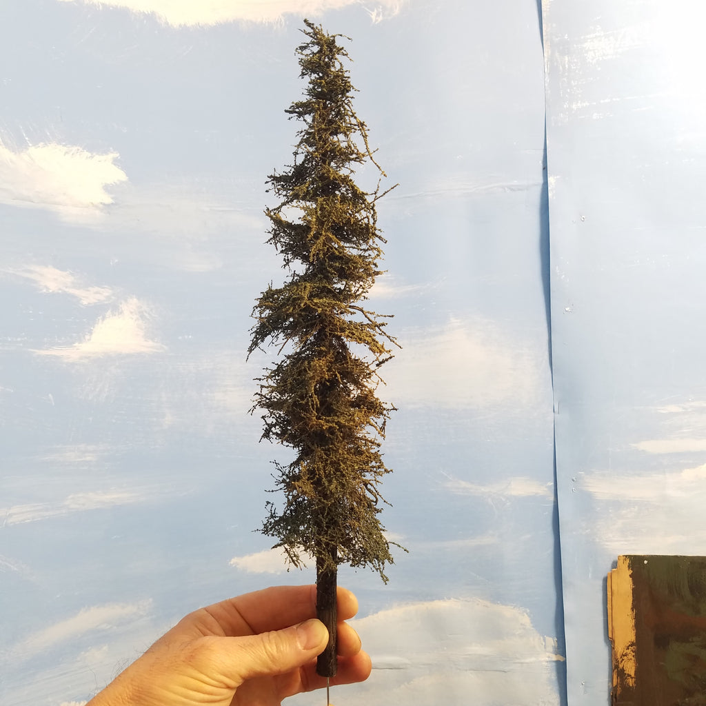 Hand holding a model of a tall evergreen Coastman's 11 inch tall fir tree against a blue sky with white clouds.