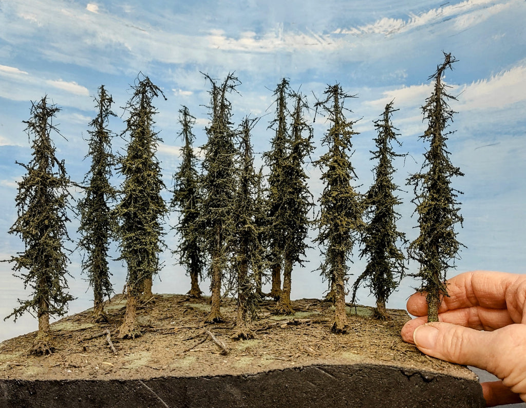 Hand holding a miniature model of a forest with trees against a blue sky.