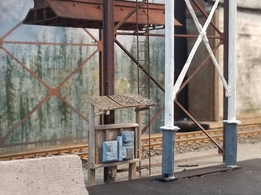 Close up of the electrical panel shack (shack not included in kit, the electrical panel is included), feeder tube support, ladder, with wood chip loader structure in the background with HO scale railroad track beneath.