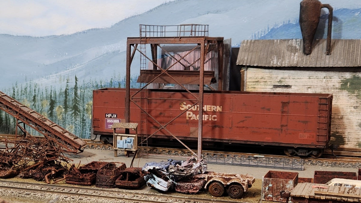 A detailed HO scale model railroad scene on Roger's Oregon Coast layout featuring a weathered Southern Pacific woodchip gondola, a trackside industrial mill structure, scrap autos, and a conveyor belt system against a backdrop of mountains and trees.
