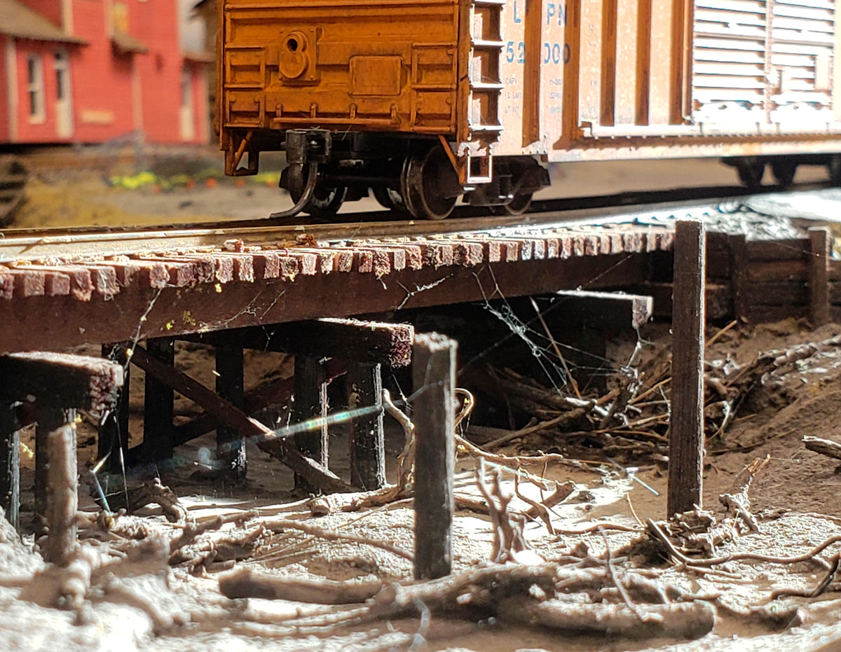 HO scale model railroad trestle with pilings taken from Coastman's Pilings product in the mud down below and a yellow boxcar sitting on the tracks above.