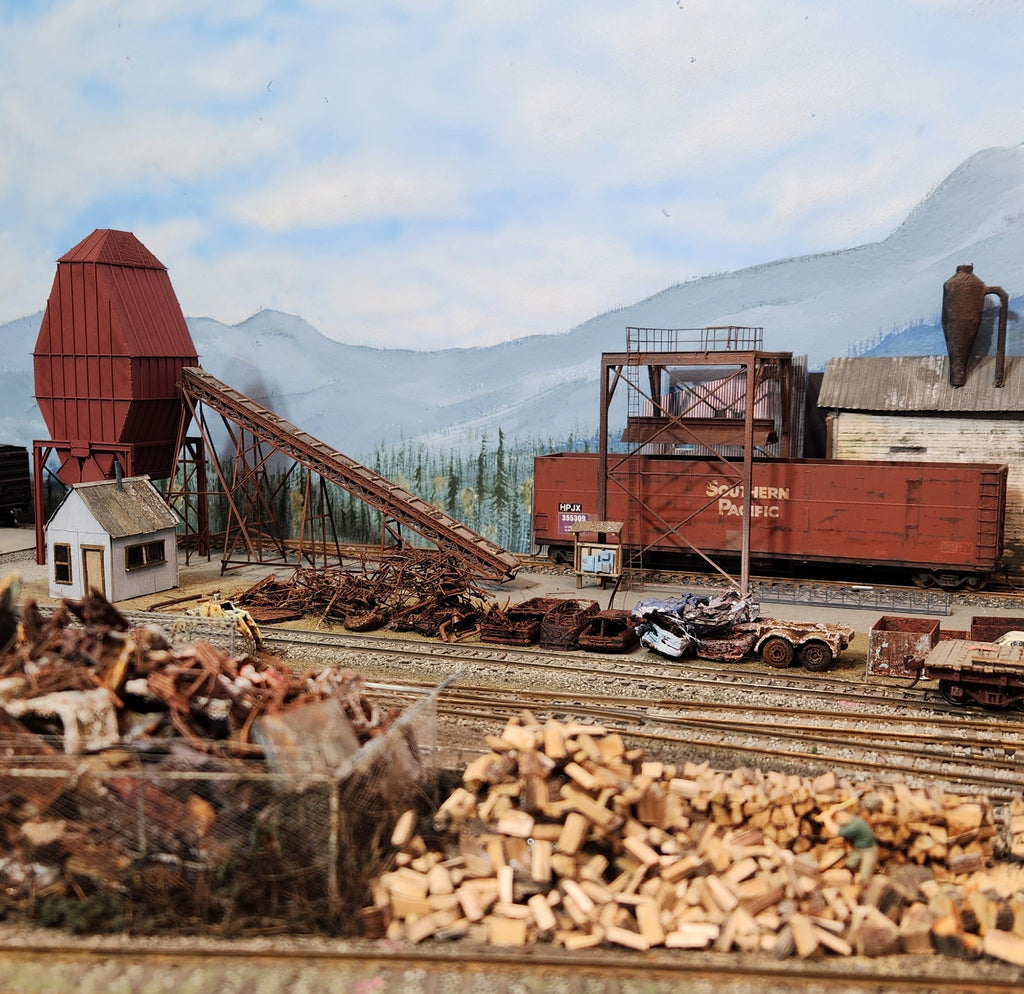 Distance photograph of HO scale wood chip loader on Roger's Oregon Coast HO Layout with HO scale wood chip gonola underneath the loader receiving wood chips and associated details including a wood chip storage bin, mill, scrap metai, and HO scale firewood scene.